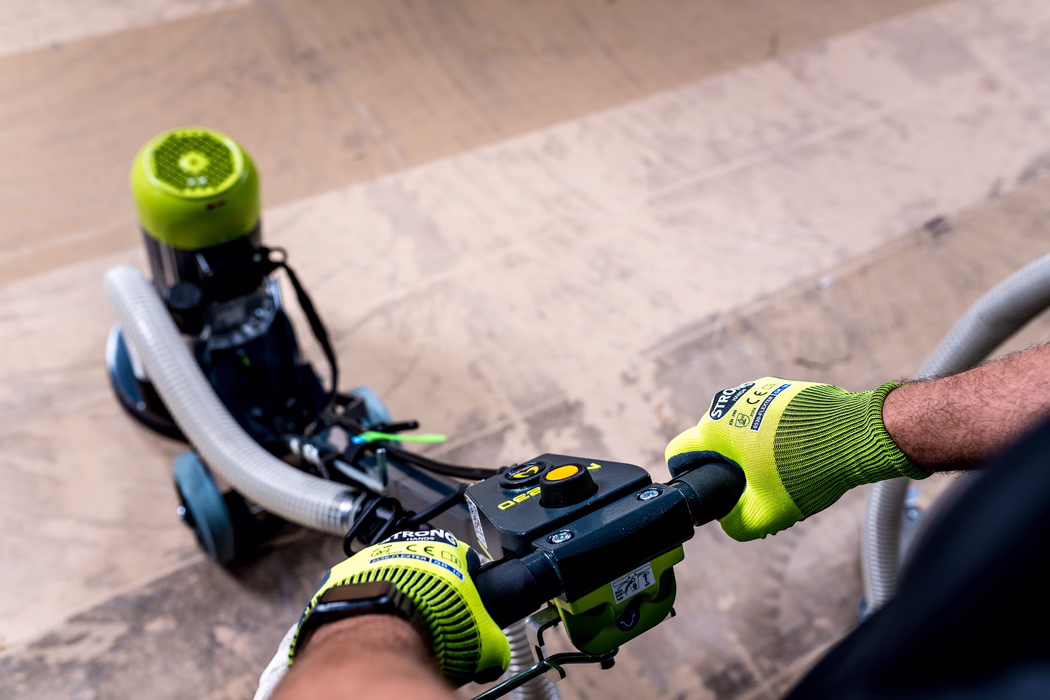 WOLFF_Application Image_Neo230_Detail14_2022-11 Close-up view of a person operating a floor sanding/polishing machine: two hands wearing high-visibility lime-green work gloves grip the black handlebar with control buttons, a flexible vacuum hose connects to the machine in the background and worn wooden floorboards are visible below. The image focuses on the equipment and hands, showing textured glove material, control dials, bolts and part of the sanding unit with wheels, capturing a workshop or renovation scene with dust and surface variations on the wood. No identifiable faces are shown.