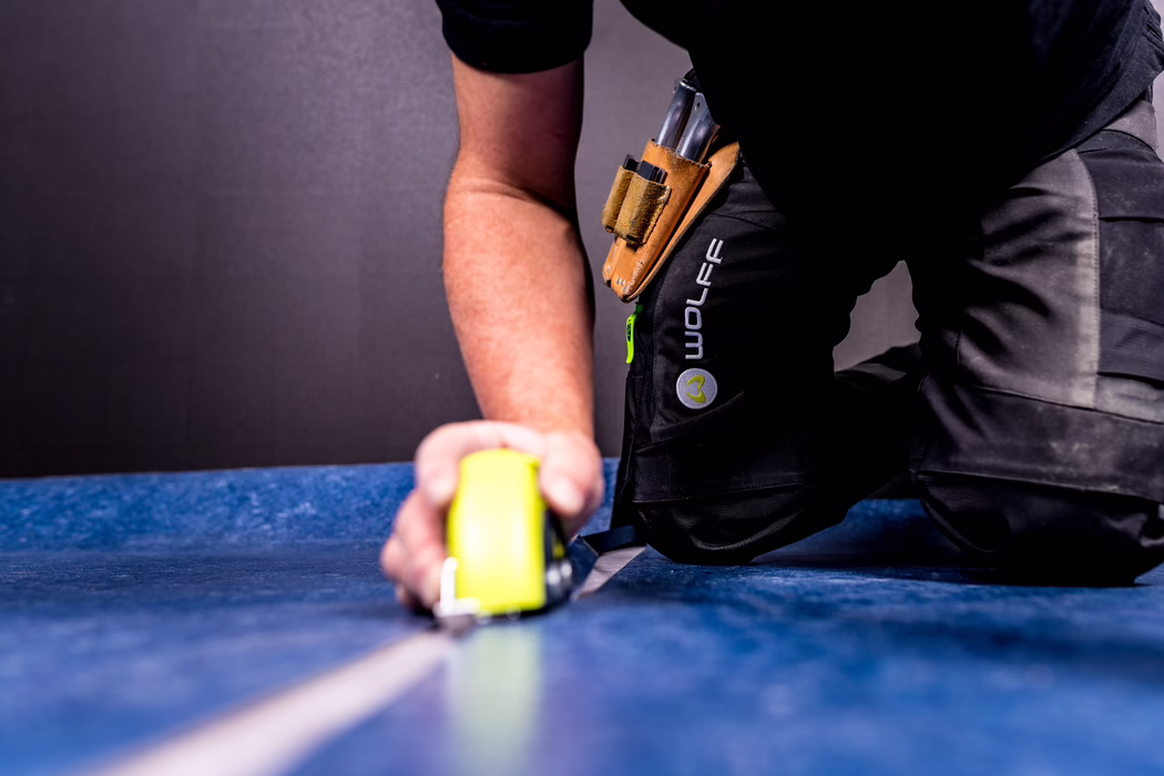 Close-up van een knielende arbeider op een blauwe ondergrond die met een gele rolmaat vooruit meet; zichtbare onderarm, leren gereedschapsriem bevestigd aan zwarte werkbroek met 'WOLFF'-logo, metalen gereedschap steekt eruit; neutrale achtergrond.