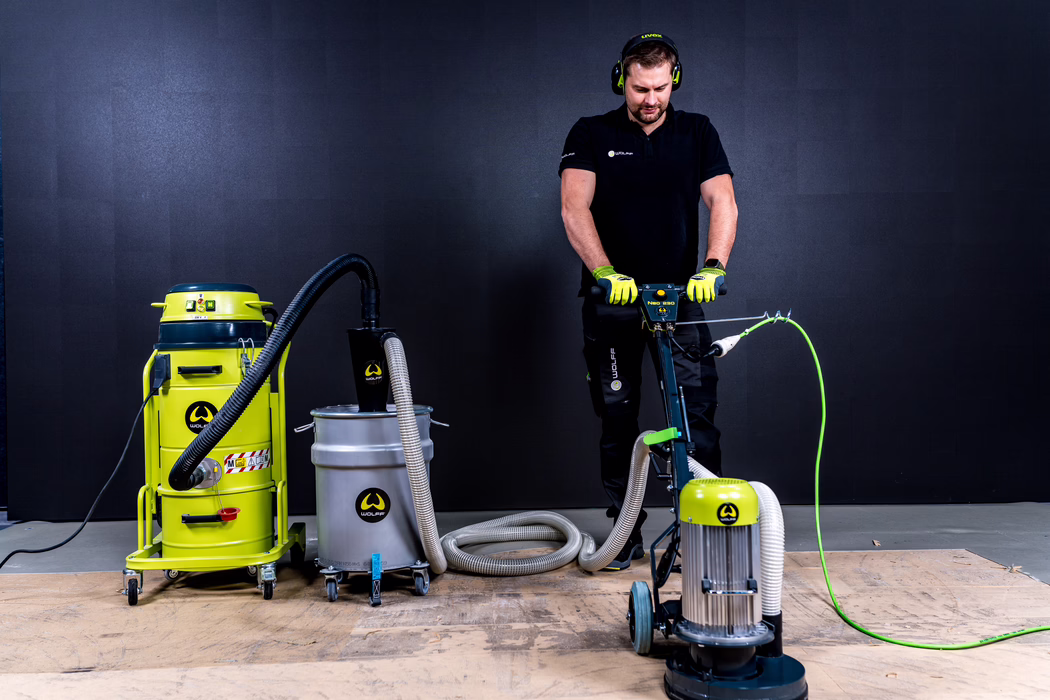WOLFF_Application Image_Neo230_Main2_2022-11 A worker operates a floor grinding machine in a studio-like setting. He stands behind a compact rotary floor grinder connected by flexible hoses to multiple industrial vacuum units and a stainless steel collection drum, all with bright lime-green accents. The man wears black work clothes, neon-green safety gloves and earmuffs and concentrates on guiding the machine over a wooden subfloor; the dark, textured wall in the background and even studio lighting emphasize the equipment, hoses and protective gear.